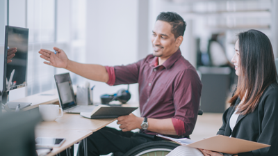 The Value of Claims Download to Agencies A man in a wheelchair sitting at a desk and showing his coworker something on the computer screen in front of them